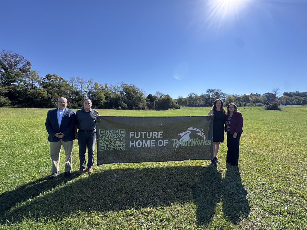 Future Facility site for MELD PrintWorks Corporation. Image shows a large field with a sign saying "future home of PrintWorks." Four people, Brian T Hamilton, Larry Vicari, Nanci Hardwick, and Brenda Rigney stand on the sides of the sign.